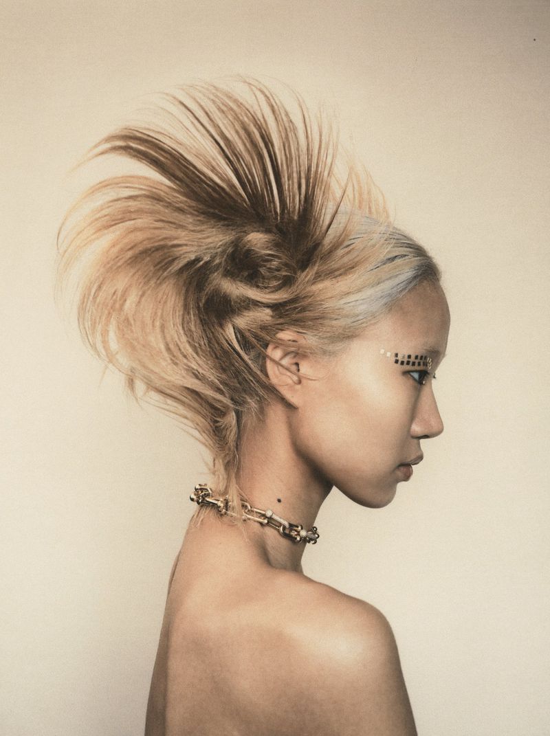 East Asian woman in profile with sculptural spiky blonde hair, graphic eye makeup, and a gold link choker against a warm neutral backdrop.