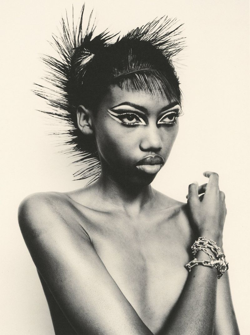 Black and white portrait of a Black woman with spiky sculpted hair and graphic winged eye makeup wearing a chunky chain bracelet.