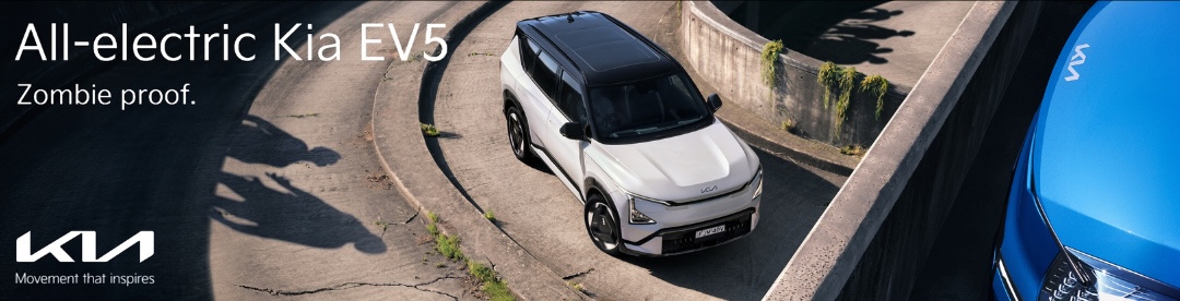 White Kia EV5 on a concrete road surrounded by walls, with blue Kia car in the foreground and shadows of people in the background.