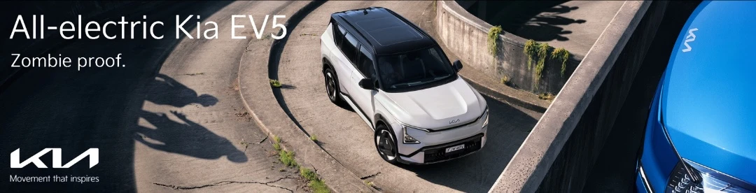White Kia EV5 on a concrete road surrounded by walls, with blue Kia car in the foreground and shadows of people in the background.