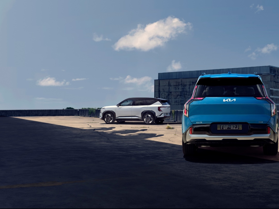 Two Kia EV9 SUVs, one teal in the foreground and one white in the background, are parked in an urban setting under a blue, cloudy sky.