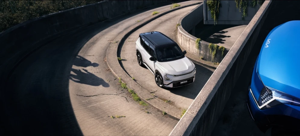 Aerial view of a white Kia EV9 SUV driving on a concrete road lined with concrete barriers, partially parked near a blue Kia.