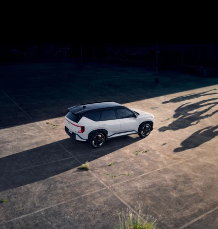 Aerial shot of a white SUV parked on a concrete surface with patches of grass and long shadows, suggesting a group of people nearby.
