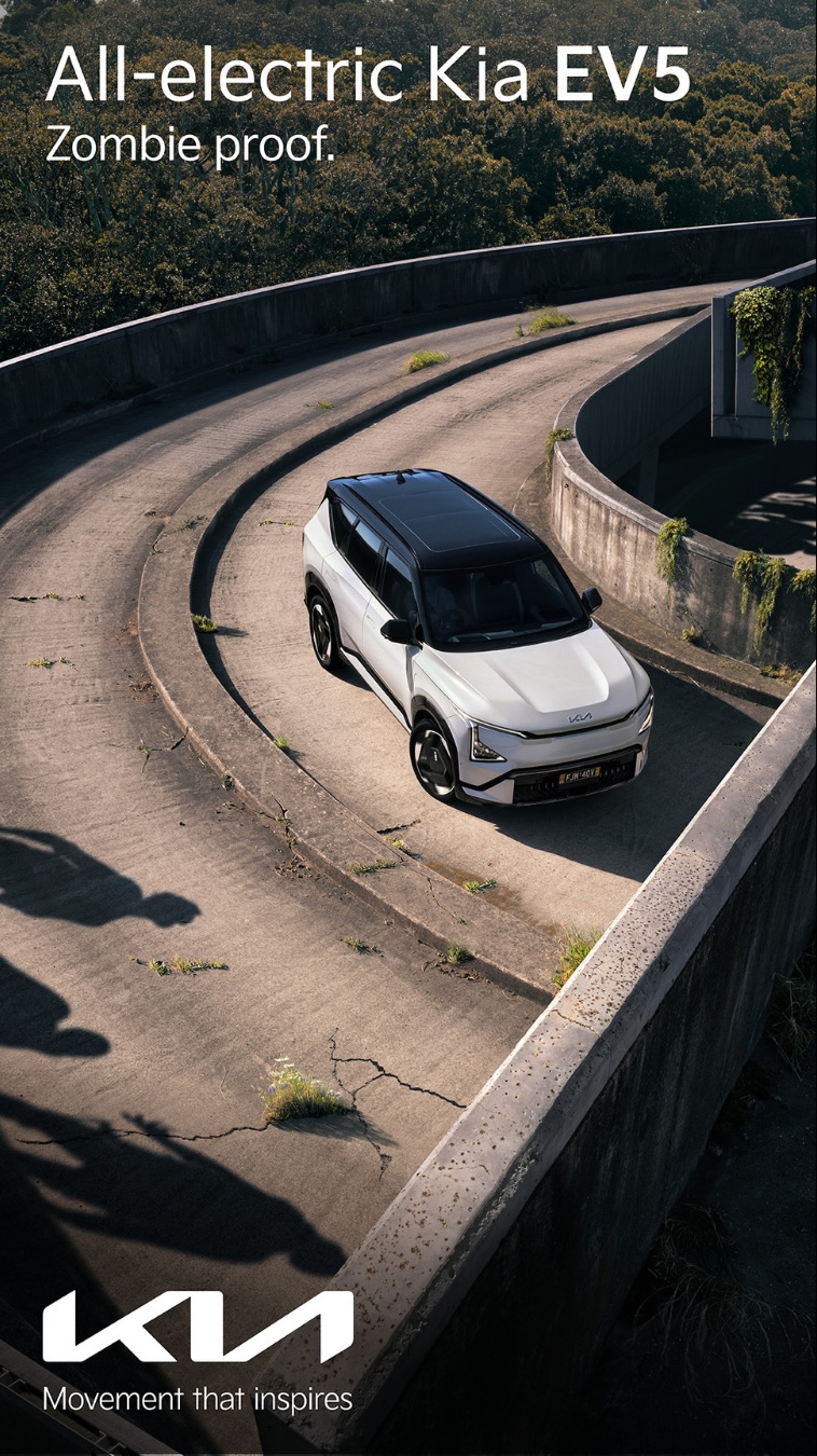 White Kia EV5 driving on a winding road with concrete barriers, surrounded by lush greenery and trees in an outdoor setting.