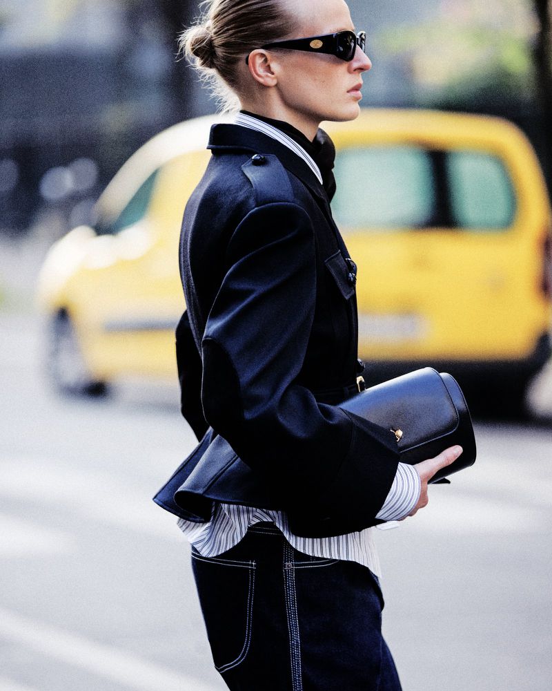 Model in dark sunglasses wearing a black jacket and carrying a handbag with a blurred yellow car in the background
