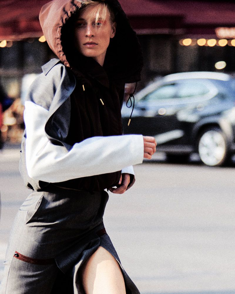 Model Anine Van Velzen walking on a city street wearing a hooded two-tone jacket and a tailored grey skirt with a slit.
