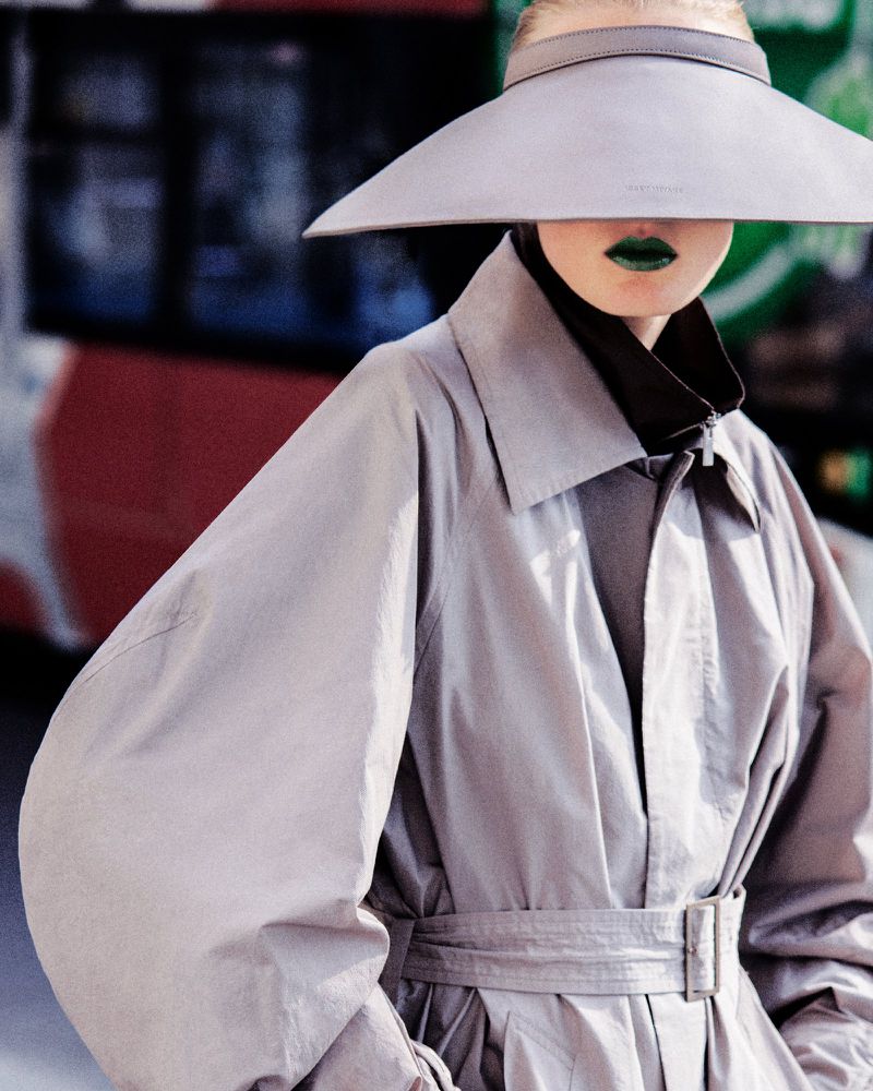 Model Anine Van Velzen wearing a wide-brim visor hat, grey belted trench coat, and green lipstick in an editorial shot.
