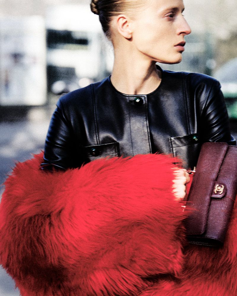 Model Anine Van Velzen wearing a black leather jacket and vivid red fur wrap while holding a burgundy clutch outdoors.