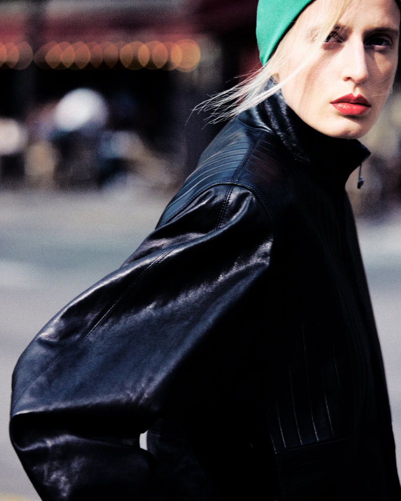 Model wearing a black leather jacket and green beanie outdoors with a blurred background.