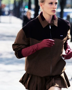 Model Anine Van Velzen running in a brown zip-up jacket, matching pleated skirt, and long burgundy gloves on an urban street.