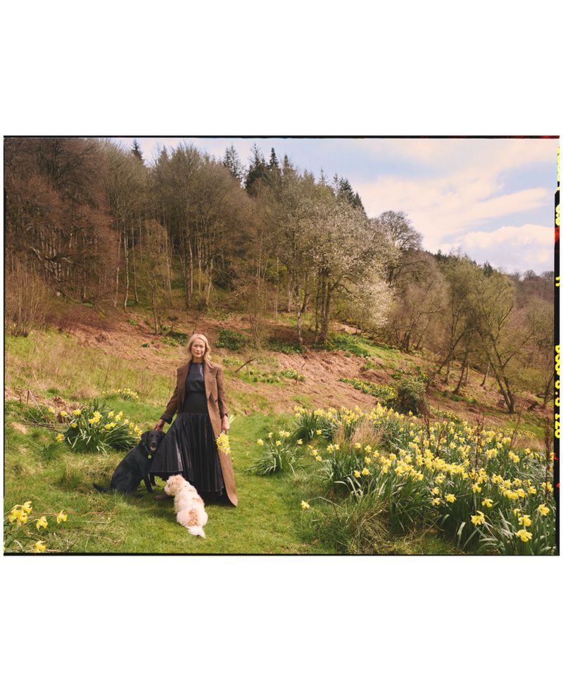 Woman in a long brown coat and black dress standing on a grassy hill with two dogs surrounded by yellow daffodils.