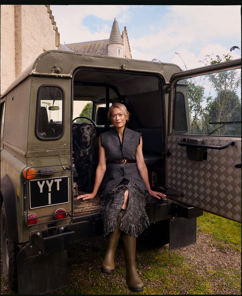 Woman in a grey tweed dress and green wellington boots seated in the back of an olive green off-road vehicle with a black dog.