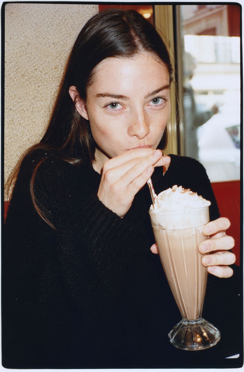 A young woman in a black knit sweater drinks a chocolate milkshake with whipped cream through a straw in a cafe booth.