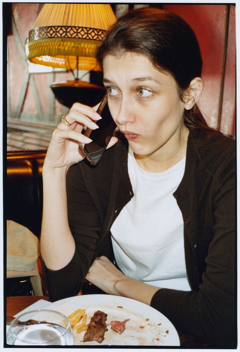 A woman talking on a smartphone seated at a restaurant table with a half-eaten meal in front of a vintage fringed table lamp.