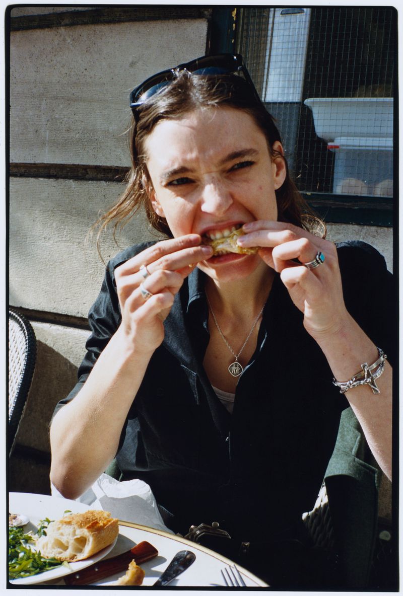 Woman with sunglasses on her head biting into crusty bread at an outdoor cafe table, wearing a black shirt and silver jewelry.