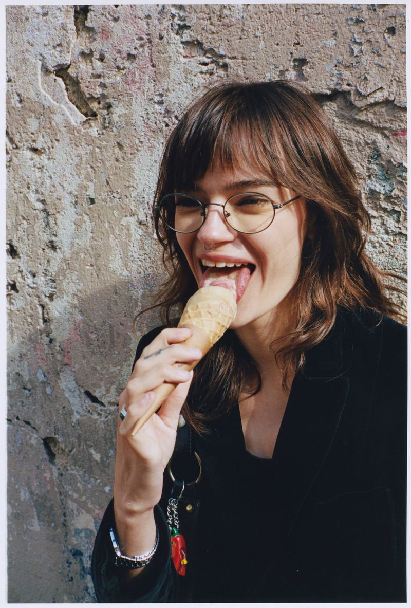 Young woman with bangs and glasses smiling while licking an ice cream cone in front of a rustic concrete wall.