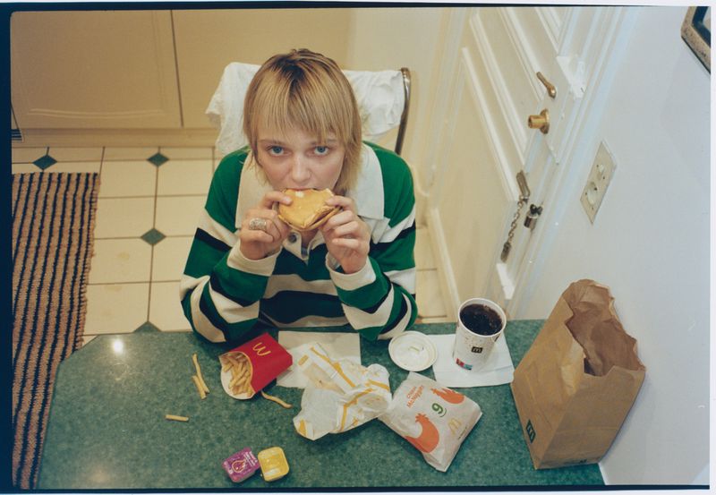 A blonde woman in a green and white striped sweater eating a fast food burger at a green kitchen counter with takeout fries and soda.