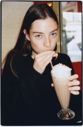 A young woman in a black knit sweater drinks a chocolate milkshake with whipped cream through a straw in a cafe booth.