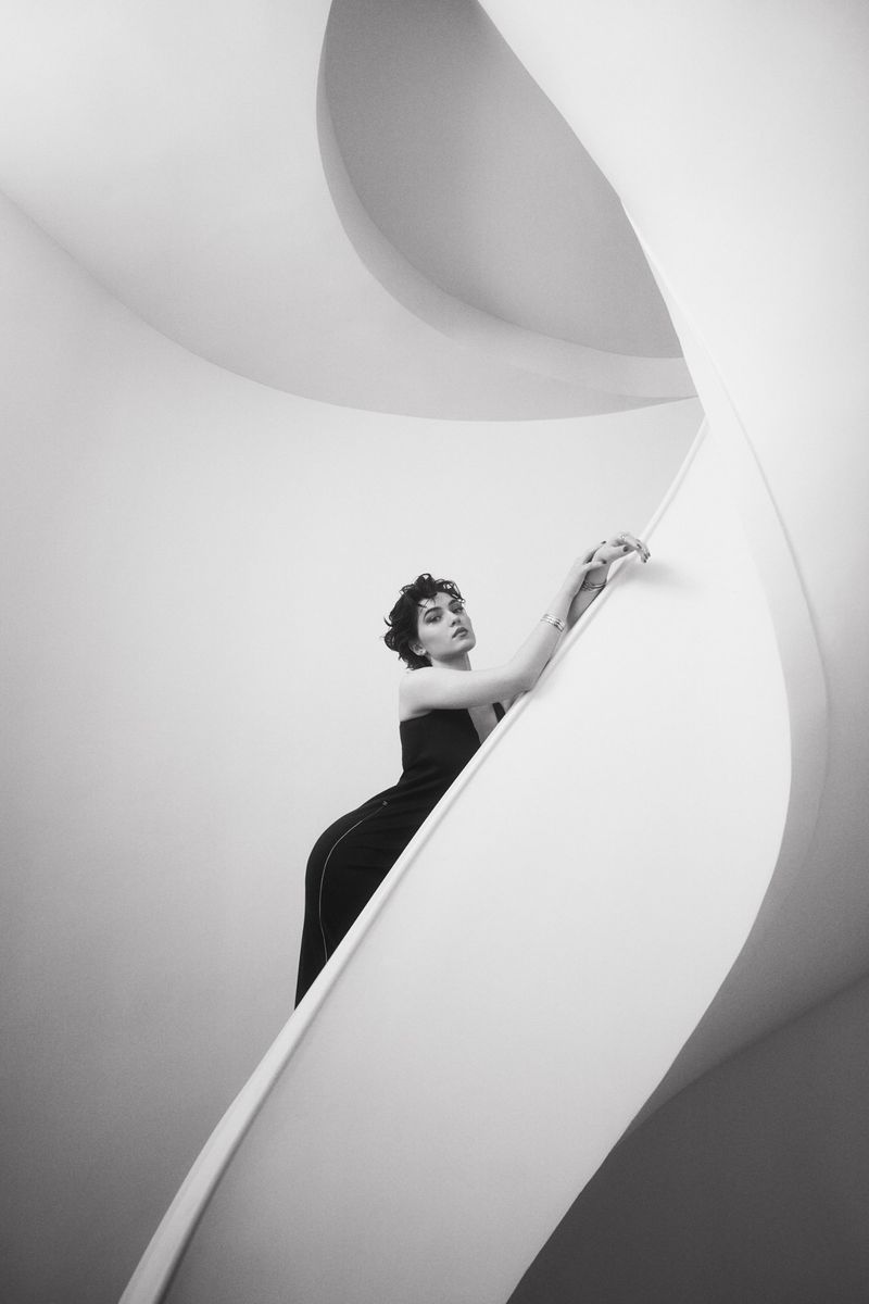 A woman in a black dress leans gracefully against the smooth sweeping curve of a white architectural spiral staircase in black and white.