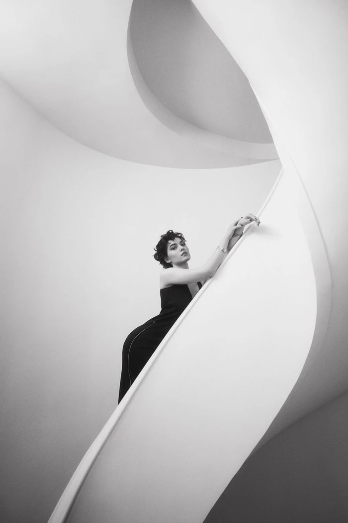 A woman in a black dress leans gracefully against the smooth sweeping curve of a white architectural spiral staircase in black and white.