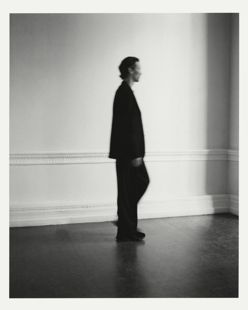 Side view of a person in a dark oversized suit walking past a white wall with classic moulding in soft light.