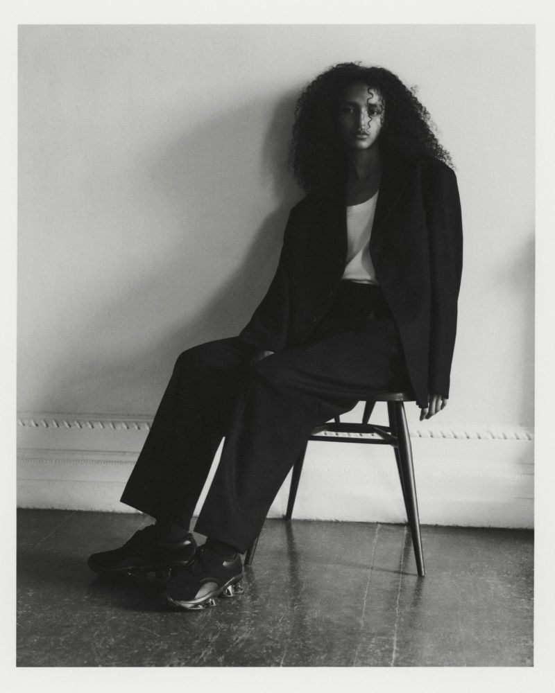 Model Tsion Teferi seated on a wooden chair wearing an oversized dark tailored suit and dark sneakers in a black and white portrait.