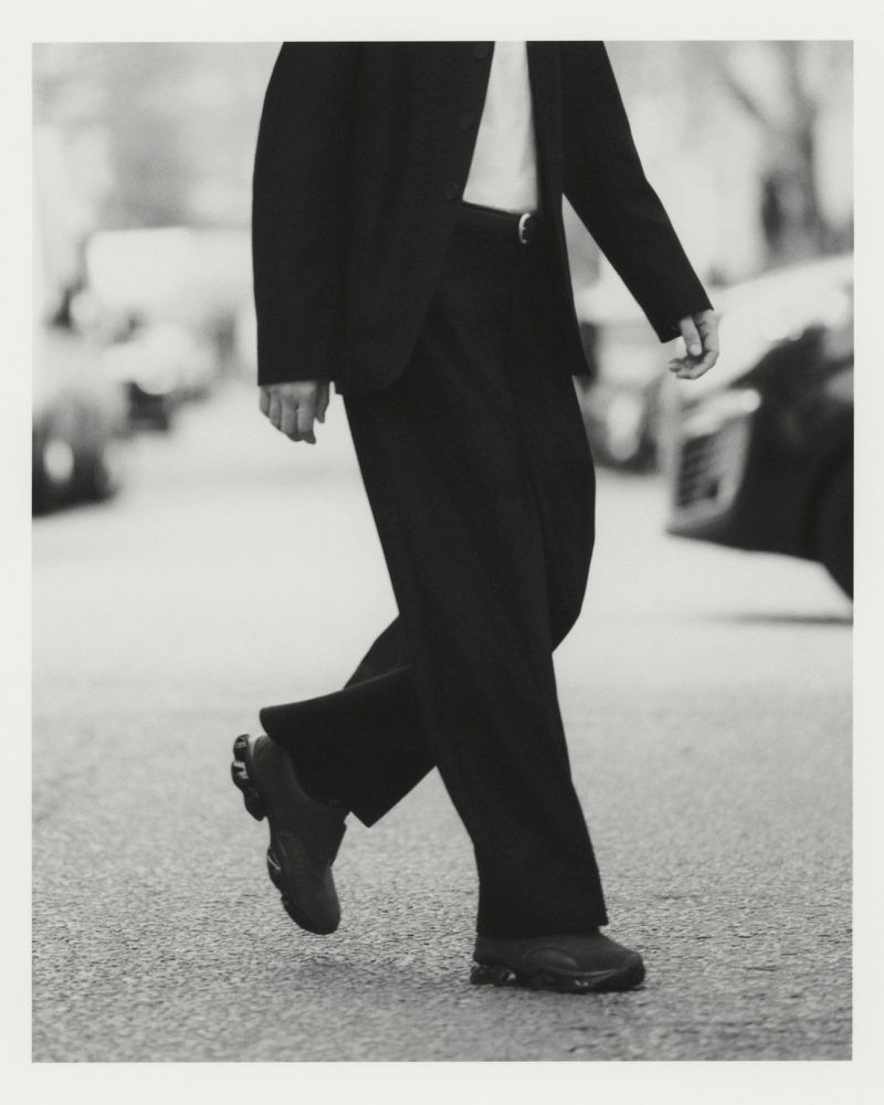 Cropped view of a model walking on a city street wearing wide-leg trousers, a tailored dark jacket, and sneakers.