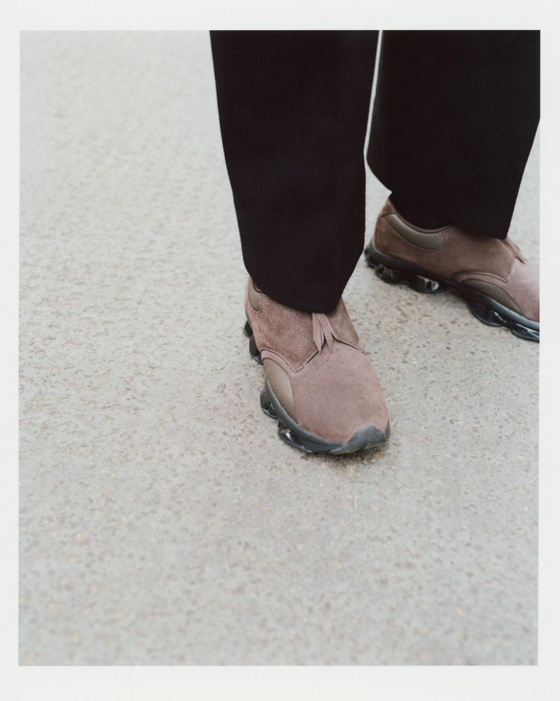 Close-up high-angle view of brown suede slip-on sneakers and black trousers standing on a textured concrete surface.