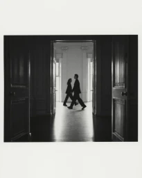 Black and white silhouette of two models walking through a bright doorway framed by dark interior doors.