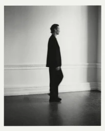 Side view of a person in a dark oversized suit walking past a white wall with classic moulding in soft light.