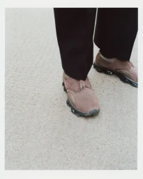 Close-up high-angle view of brown suede slip-on sneakers and black trousers standing on a textured concrete surface.
