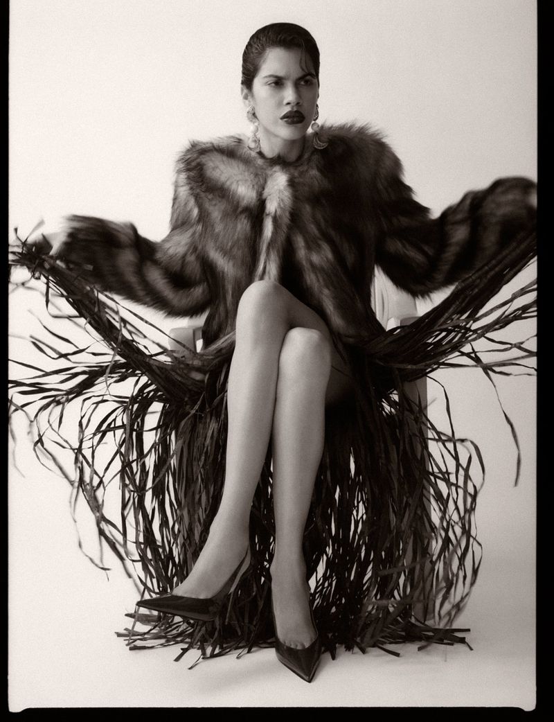 Model Valeria Gomez seated in a fur jacket and shredded fringe skirt with crossed legs against a white studio background.