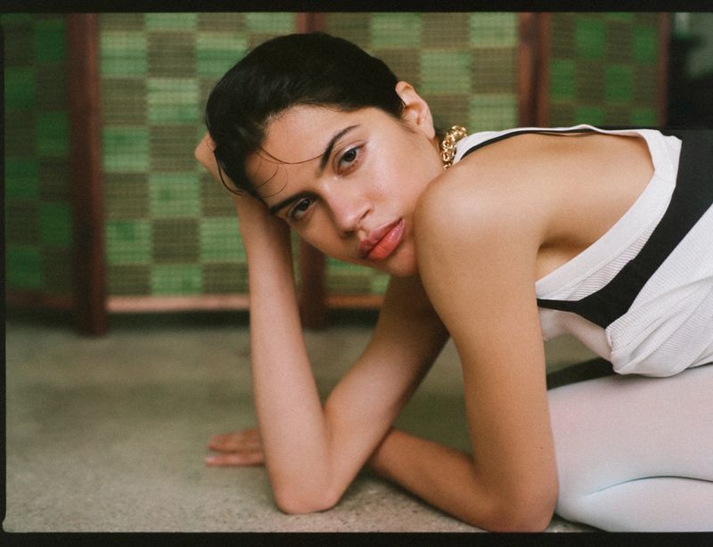 Model Valeria Gomez crouching on the floor wearing a white top and gold chain necklace in front of a green woven screen.