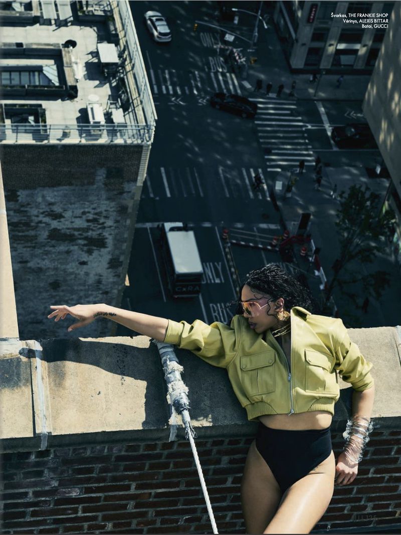 Fashion model in a cropped chartreuse jacket and black bottoms leaning over a brick rooftop overlooking a city street.
