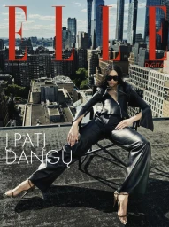 Model in a black leather outfit and sunglasses seated on a folding chair on a rooftop with a city skyline behind her.