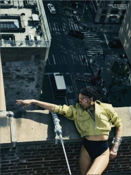 Fashion model in a cropped chartreuse jacket and black bottoms leaning over a brick rooftop overlooking a city street.