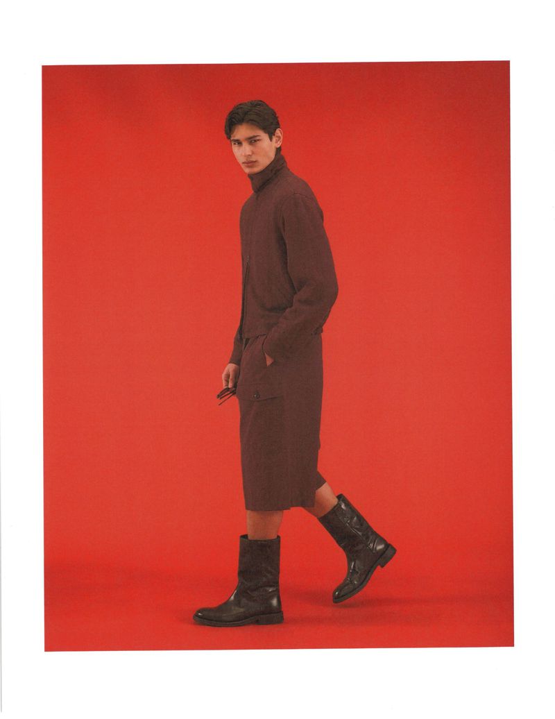 Model Ellis Mueller wears a brown jacket, matching shorts, and black leather boots against a solid red studio background.