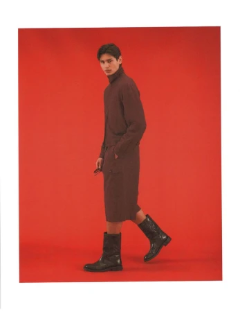 Model Ellis Mueller wears a brown jacket, matching shorts, and black leather boots against a solid red studio background.