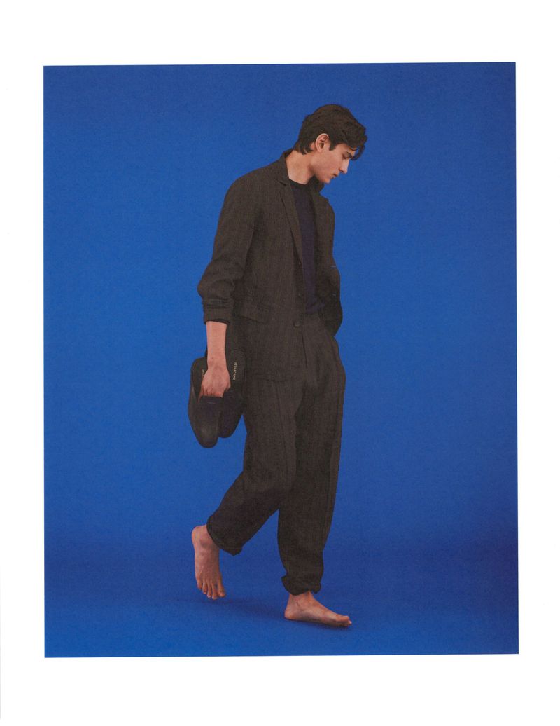 Model Ellis Mueller walking barefoot while holding black shoes in a dark suit against a vibrant blue studio backdrop.