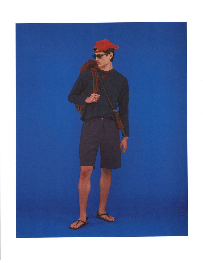 Model Ellis Mueller wears a navy long-sleeve shirt, dark shorts, sandals, and a red cap against a bold blue studio backdrop.