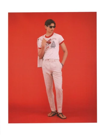 Model wearing dark sunglasses, graphic t-shirt, and light trousers standing against a solid red background for American GQ Style