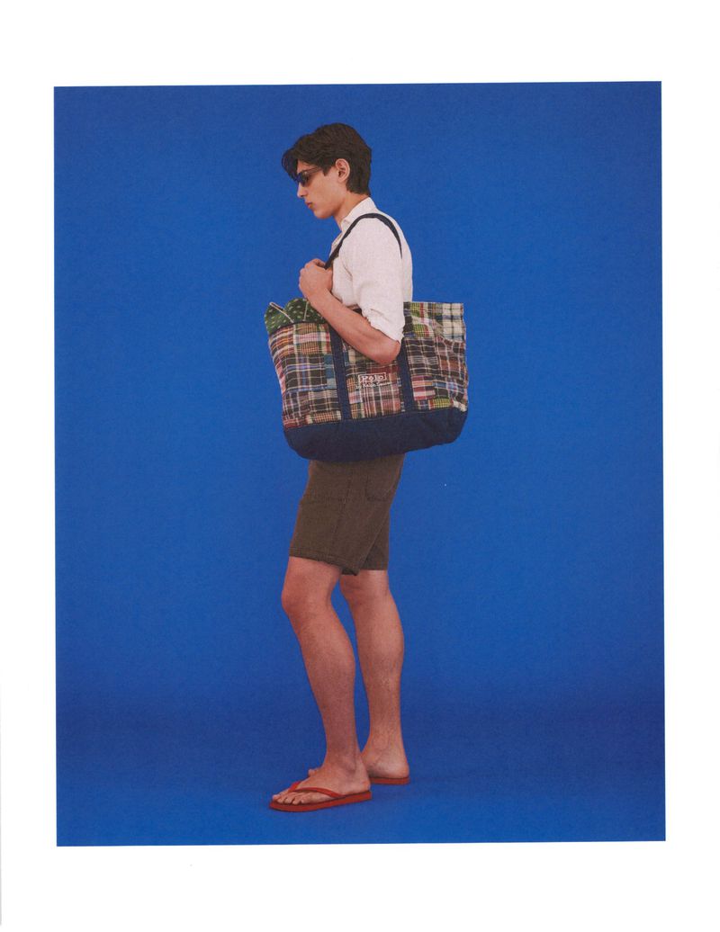 Model Ellis Mueller standing in profile with a patchwork plaid tote bag, white shirt, shorts, and flip-flops against a blue background.