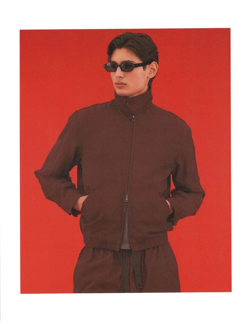 Ellis Mueller wearing a dark brown zip jacket and black sunglasses with hands in pockets against a bright red studio background.