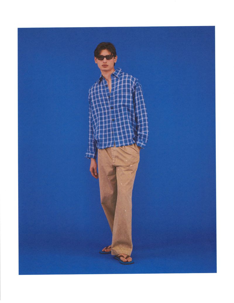 Model wearing dark sunglasses, a blue plaid button-down shirt, and beige trousers against a vibrant blue studio backdrop.