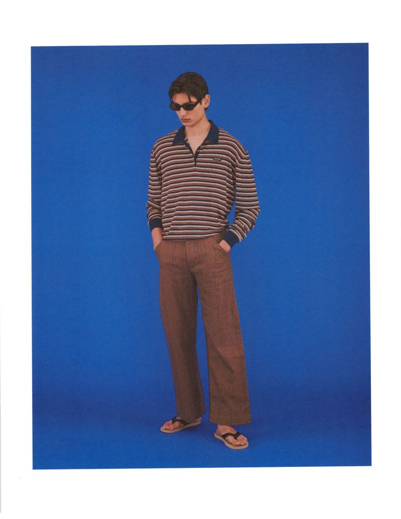 Model Ellis Mueller standing in sunglasses, a striped collared long-sleeve shirt, and brown trousers against a solid blue backdrop.