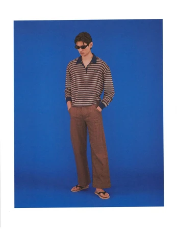 Model Ellis Mueller standing in sunglasses, a striped collared long-sleeve shirt, and brown trousers against a solid blue backdrop.