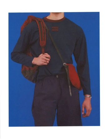 Cropped shot of a model wearing a navy blue long-sleeve shirt and dark trousers with a burgundy backpack against a blue backdrop.