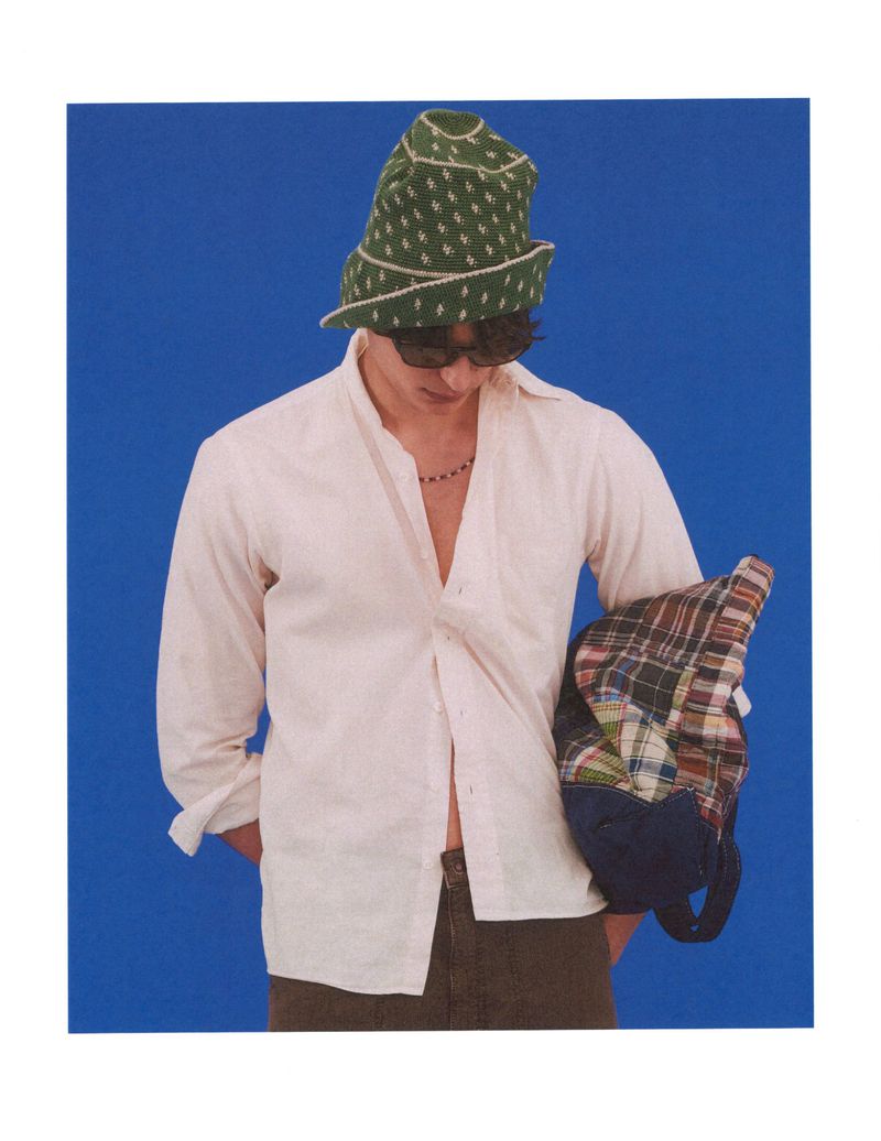 Model Ellis Mueller wearing a relaxed white open shirt, green patterned bucket hat, and sunglasses against a blue backdrop.