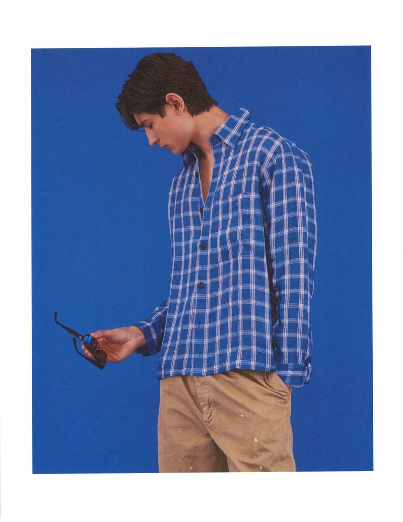 Model Ellis Mueller wearing a blue plaid shirt holding sunglasses against a solid blue seamless backdrop for American GQ Style.
