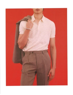 Model Ellis Mueller in a cream polo shirt and taupe pleated trousers holding a blazer over his shoulder on a bright red backdrop.